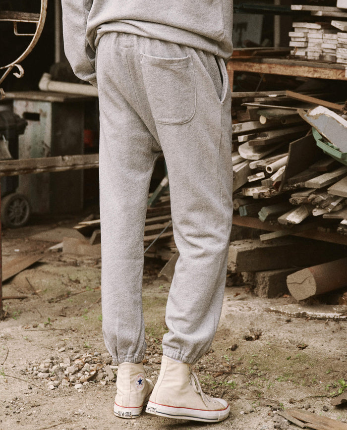 A person dressed in The Men's Stadium Sweatpant in Varsity Grey by THE GREAT. HOL 21 MENS, paired with white high-top sneakers, stands in a cluttered, rustic environment with stacks of wood in the background.