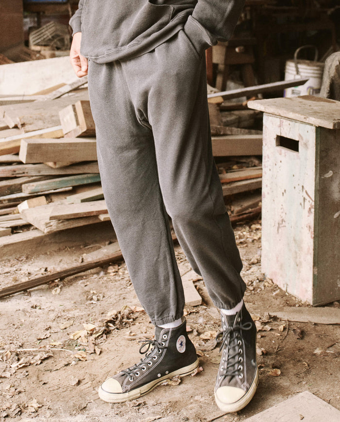 A person standing in a workshop is wearing THE GREAT. HOL 21 MENS' The Men's Stadium Sweatpant in Washed Black, paired with black high-top sneakers and white socks. The background includes wooden planks and a green workbench, creating a scene that exudes vintage athleticwear vibes, perfect for the laid-back lifestyle of Los Angeles.