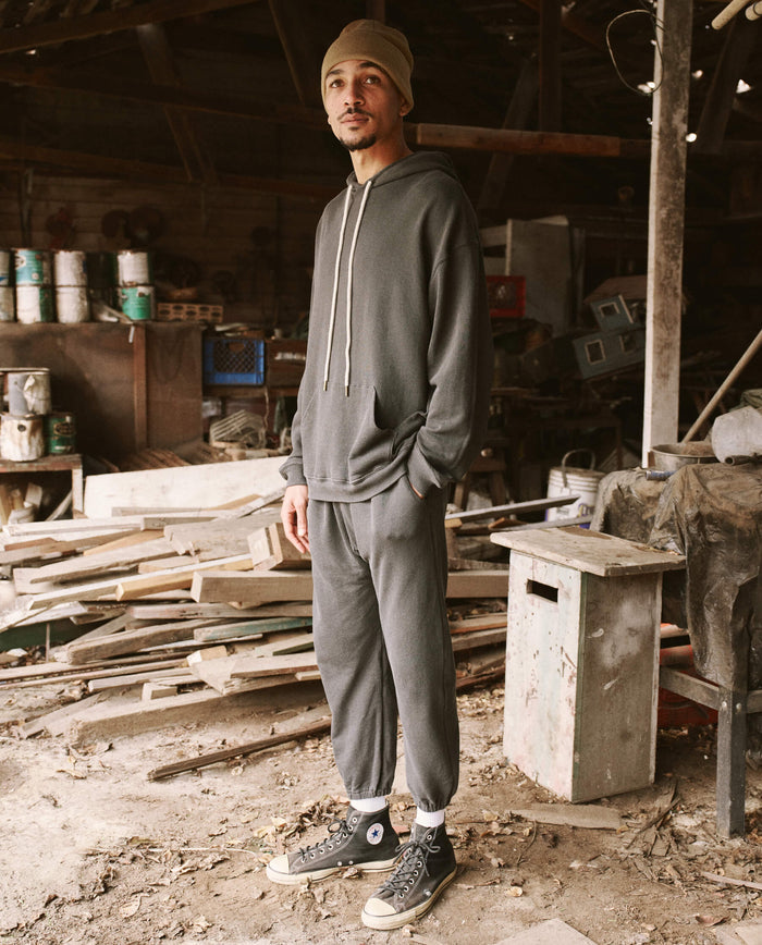 A person stands in a Los Angeles workshop wearing a beanie, hoodie, and vintage athleticwear including The Men's Stadium Sweatpant in Washed Black by THE GREAT. HOL 21 MENS, with hands in pockets. The workshop has scattered wooden planks, paint cans, and various tools.