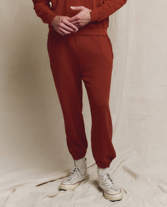 A man in THE GREAT. F25 MENS The Men's Stadium Sweatpant in Brick and matching sweatshirt pairs the retro look with white high-top sneakers, standing before a neutral fabric backdrop for vintage athletic style.