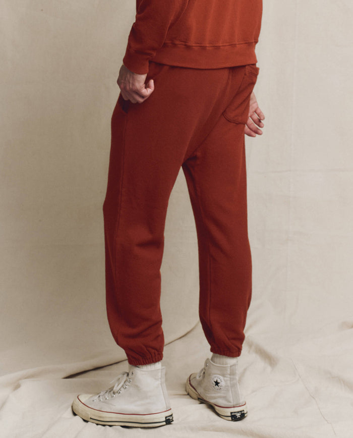 A person wears THE GREAT. F25 MENS The Men's Stadium Sweatpant in Solid Brick and a matching top, standing on a light fabric surface with white high-top sneakers—evoking vintage athletic wear and classic Los Angeles knits.