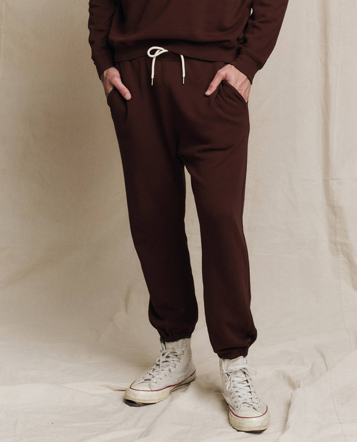 Model wears THE GREAT. F25 MENS The Men's Stadium Sweatpant in Driftwood, paired with a matching top and white high-top sneakers, standing hands-in-pockets against a neutral backdrop.