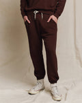 Model wears THE GREAT. F25 MENS The Men's Stadium Sweatpant in Driftwood, paired with a matching top and white high-top sneakers, standing hands-in-pockets against a neutral backdrop.