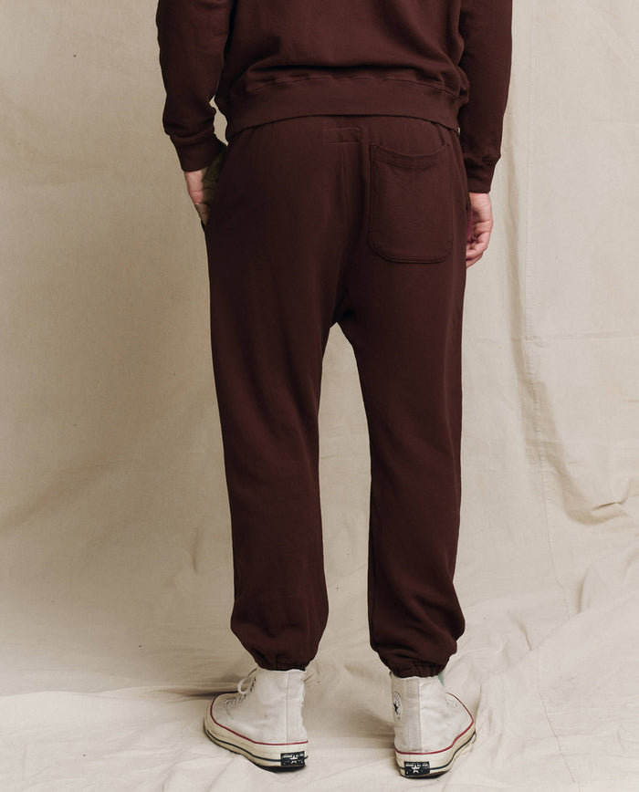 A person wearing THE GREAT. F25 MENS The Men's Stadium Sweatpant. Solid -- Driftwood stands with their back to a beige backdrop, paired with white high-top sneakers.