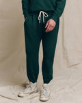 A person wearing The Men's Stadium Sweatpant in Solid Pinyon from THE GREAT. HOL 24 MEN and white high-top sneakers stands with hands in pockets, framed against a plain beige fabric backdrop.