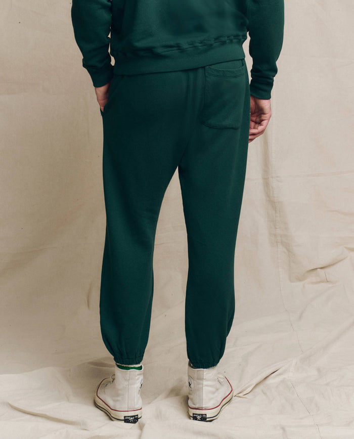 A person stands facing backward, wearing THE GREAT. HOL 24 MEN's The Men's Stadium Sweatpant in Pinyon, a matching top, and white sneakers against a beige backdrop.