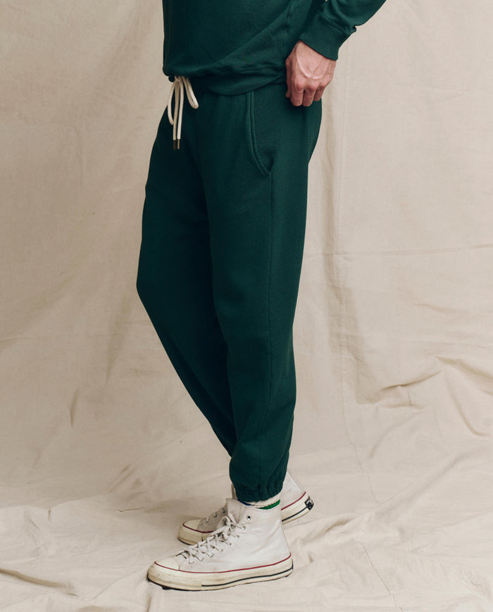 A person wearing The Men's Stadium Sweatpant in Pinyon by THE GREAT. HOL 24 MEN and white high-top sneakers stands on a beige fabric backdrop.