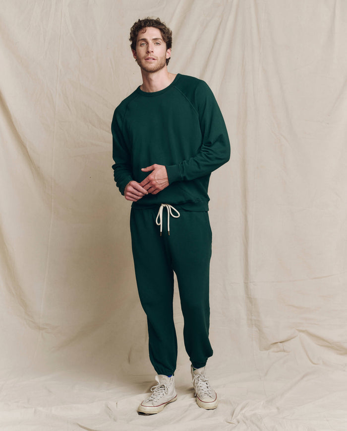 A person stands against a beige backdrop wearing The Men's Stadium Sweatpant in Pinyon by THE GREAT. HOL 24 MEN, paired with white sneakers.