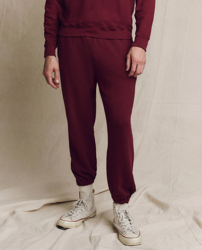 A person stands on a cream fabric backdrop, showcasing the lower body clad in THE GREAT. HOL 24 MEN's The Men's Stadium Sweatpant in Plum and white sneakers.