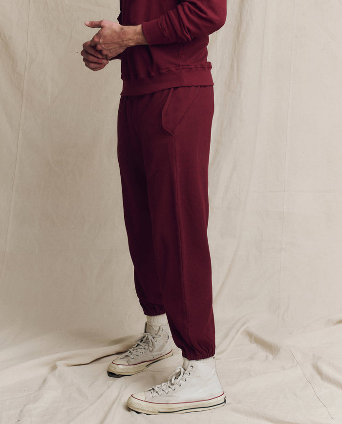 A person wearing The Men's Stadium Sweatpant in Plum from THE GREAT. HOL 24 MEN, paired with white sneakers, epitomizes men's activewear style as they stand against a beige backdrop.