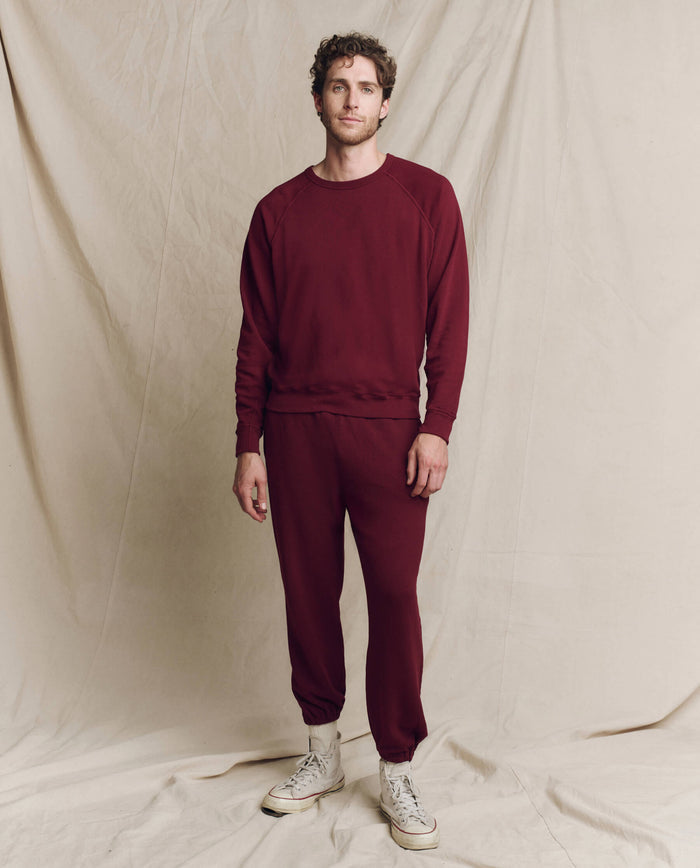 A person in THE GREAT. HOL 24 MEN's The Men's Stadium Sweatpant in Solid -- Plum stands on a beige fabric backdrop, wearing white sneakers.