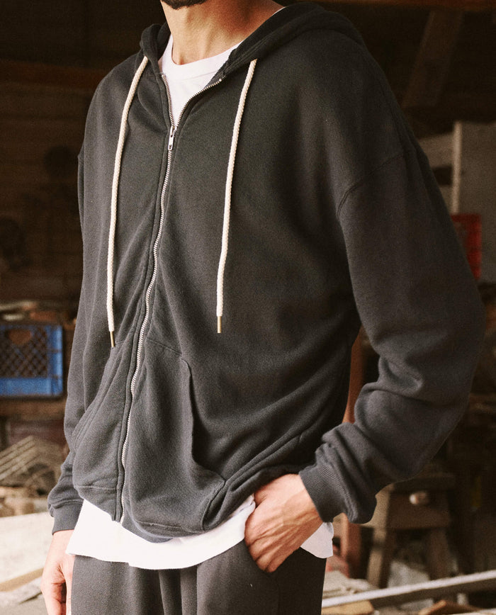 A person stands with one hand in their pocket, wearing THE GREAT. SP22 D2 MENS The Men's Zip Up Hoodie in Almost Black over a white shirt in a casually lit indoor setting.