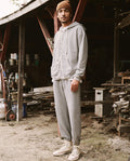 A person in The Men’s Zip Up Hoodie -- Varsity Grey from THE GREAT. SP22 D2 MENS and sweatpants stands outside an old wooden shed. They are wearing a brown beanie and white sneakers. Various tools and materials are scattered around the shed, highlighting the versatile nature of their attire.
