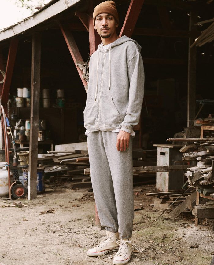A person in The Men’s Zip Up Hoodie -- Varsity Grey from THE GREAT. SP22 D2 MENS and sweatpants stands outside an old wooden shed. They are wearing a brown beanie and white sneakers. Various tools and materials are scattered around the shed, highlighting the versatile nature of their attire.