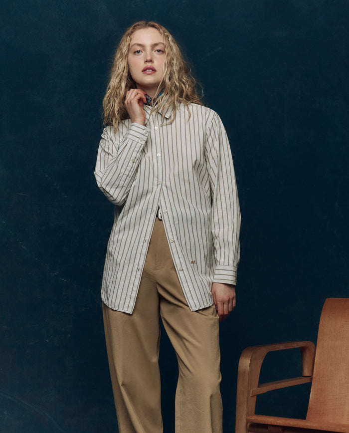 A person with long, wavy blonde hair stands beside a wooden chair against a dark background, wearing THE GREAT. PS26 COLLECTORS' The Menswear Button Up in Ivy League Stripe and beige pants.