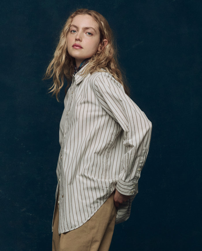 A person with long, wavy blonde hair wears THE GREAT. PS26 COLLECTORS The Menswear Button Up. -- Ivy League Stripe with white pinstripes and tan pants, standing against a dark background and glancing over their shoulder.