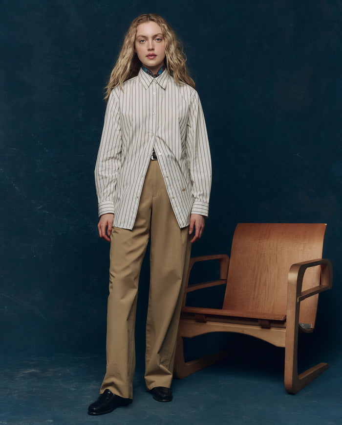 A person with long blonde hair stands before a wooden chair, wearing THE GREAT. PS26 COLLECTORS The Menswear Button Up in Ivy League Stripe, beige trousers, and black shoes against a dark background.