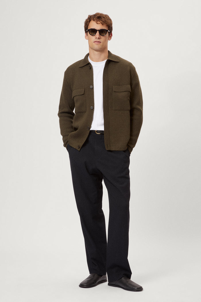 Man wearing a merino green jacket, white t-shirt, black pants, and sunglasses on a white background