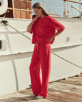 A woman stands on a dock by a white boat, wearing THE GREAT. SU26 The Micro Terry Horseshoe Sweatpant in Pimento with a matching top, gazing down and touching her face.
