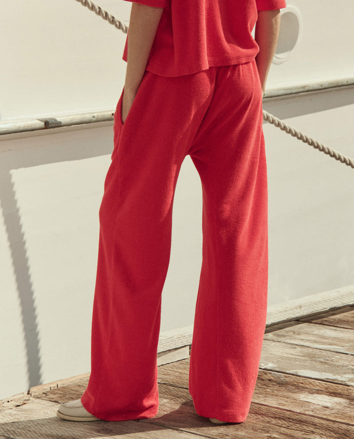 A person stands on a wooden dock near a white boat, hands in pockets, wearing THE GREAT. SU26's The Micro Terry Horseshoe Sweatpant in Pimento with a matching top.