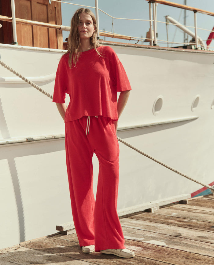 A person in THE GREAT. SU26 The Micro Terry Horseshoe Sweatpant in Pimento stands on a wooden dock beside a white boat.