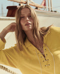 A person with long hair poses outdoors near a sailboat, wearing THE GREAT. SU26 Micro Terry Lace Up Tunic in Margarita and leaning on one arm.