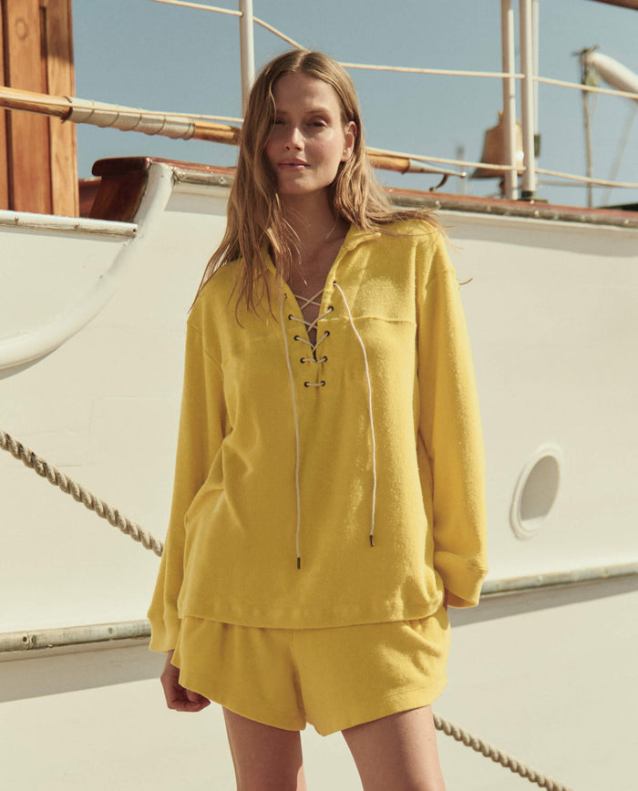 A person stands on a boat wearing THE GREAT. SU26 Micro Terry Lace Up Tunic in Margarita with matching shorts, sunlight casting natural shadows.