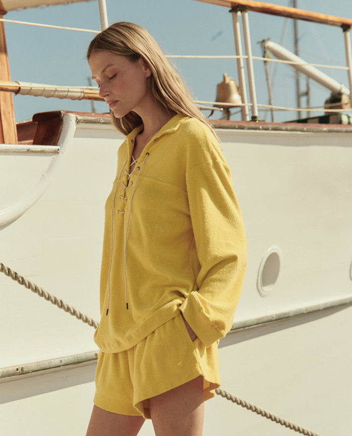 A woman wearing THE GREAT. SU26 The Micro Terry Lace Up Tunic in Margarita stands next to a white boat, her head slightly down and eyes closed.