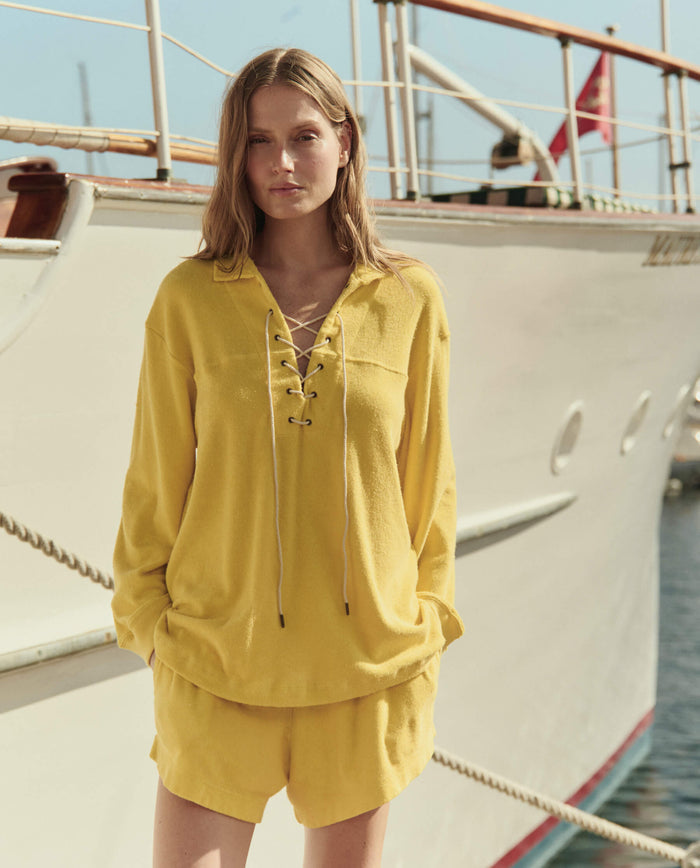 A woman wears THE GREAT. SU26 The Micro Terry Lace Up Tunic in Margarita as she stands in front of a white boat by the water.