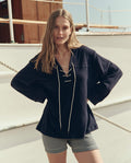 A woman stands with hands on hips on a boat deck in daylight, wearing THE GREAT. SU26 The Micro Terry Lace Up Tunic in navy and striped shorts.
