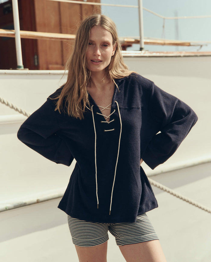 A woman stands with hands on hips on a boat deck in daylight, wearing THE GREAT. SU26 The Micro Terry Lace Up Tunic in navy and striped shorts.