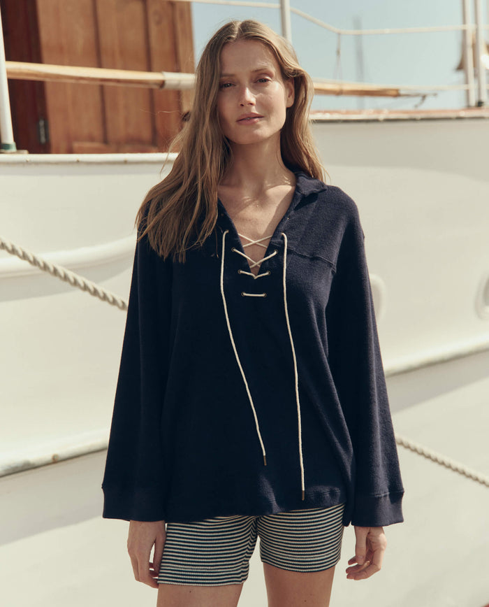 A woman stands outdoors in front of a white boat wearing THE GREAT. SU26 The Micro Terry Lace Up Tunic in navy and striped shorts.