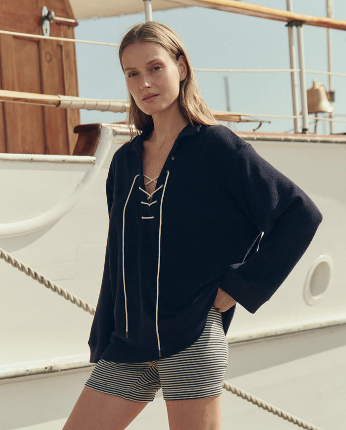 A person stands on the deck of a boat, wearing THE GREAT. SU26's Micro Terry Lace Up Tunic in navy and striped shorts.