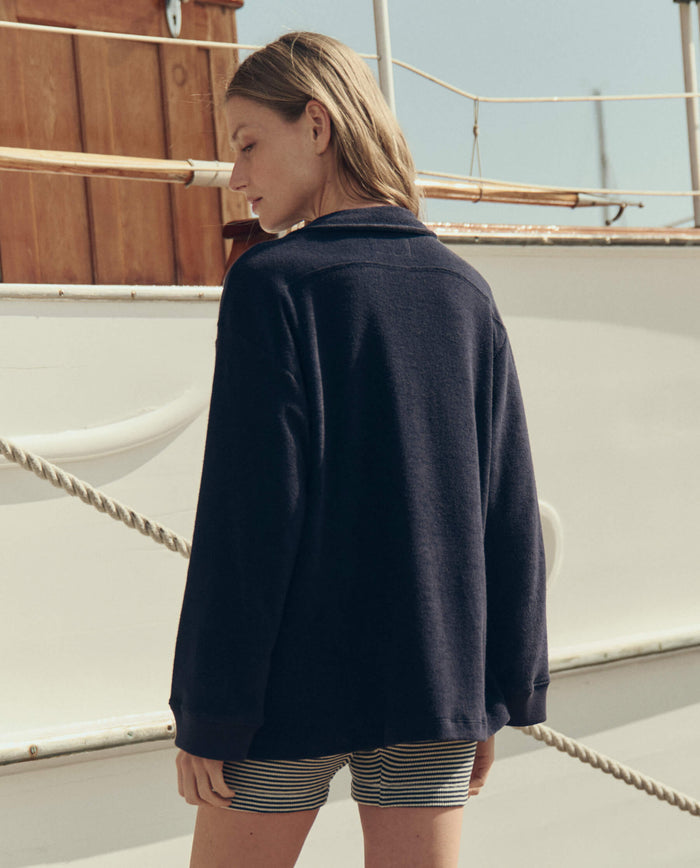 A person stands outdoors with their back to the camera, wearing THE GREAT. SU26 The Micro Terry Lace Up Tunic in Navy, near a white boat with wooden details and rope railing.
