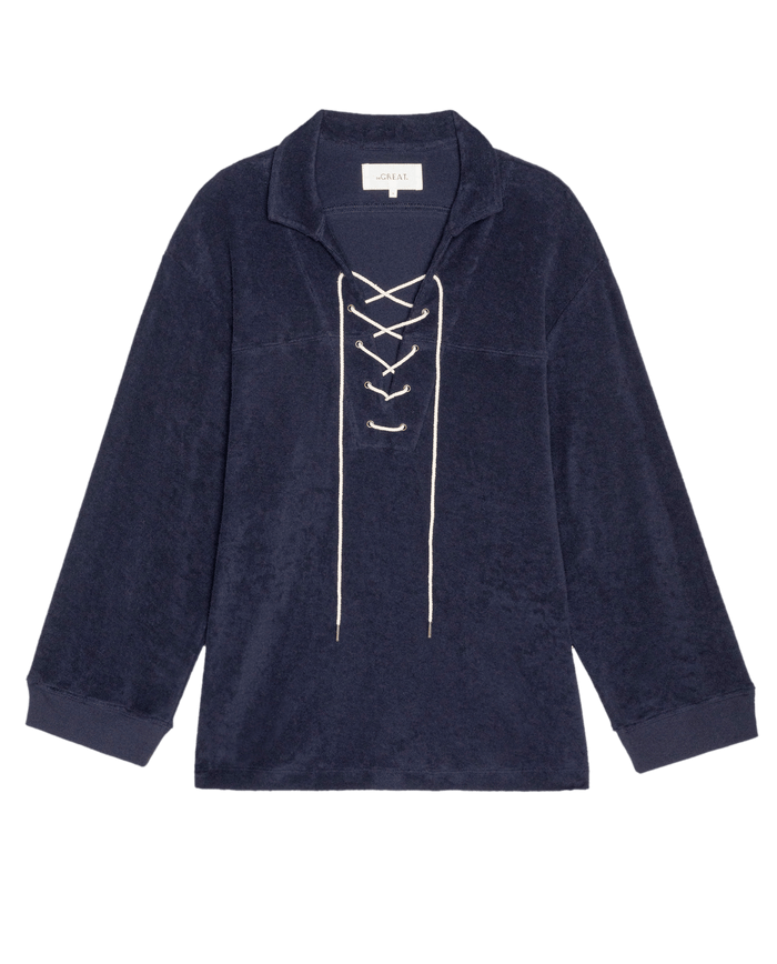 The Micro Terry Lace Up Tunic by THE GREAT. SU26 features a navy blue color, long sleeves, a collar, and white lace-up front detail in soft micro terry fabric, shown on a plain white background.