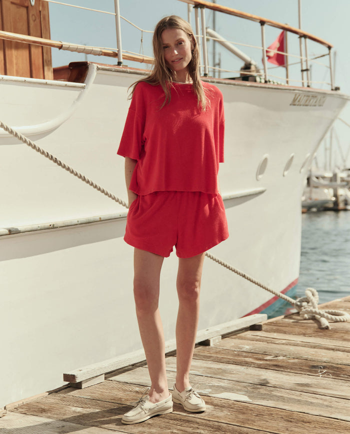 A woman stands on a dock beside a white boat, wearing THE GREAT. SU26's The Micro Terry Swing Short in Pimento and a matching red top, with one hand in her pocket and the other by her side.