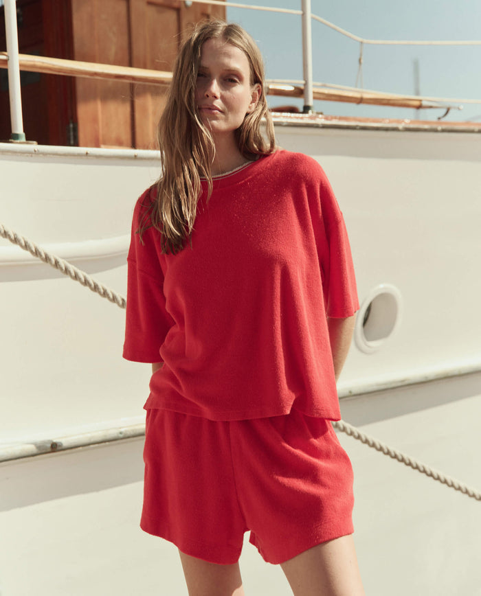 A woman in THE GREAT. SU26 The Micro Terry Swing Short in Pimento stands before a white boat, styled with a loose-fitting matching red short-sleeve top.