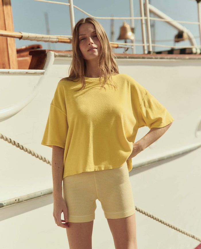 A woman stands outdoors in front of a boat, wearing THE GREAT. SU26 The Relay Short in Soft Yellow Stripe and a loose yellow t-shirt, with one hand on her hip.