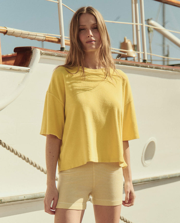 A woman stands before a boat, wearing THE GREAT. SU26’s The Micro Terry Track Crew in Margarita and light yellow striped shorts, sunlight casting shadows.