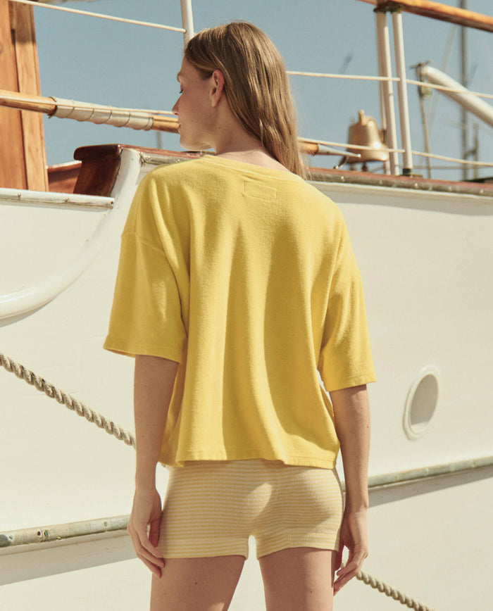 A woman with long hair stands outdoors facing a docked boat, wearing THE GREAT. SU26 The Micro Terry Track Crew in Margarita and light yellow shorts, capturing a relaxed track crew vibe.