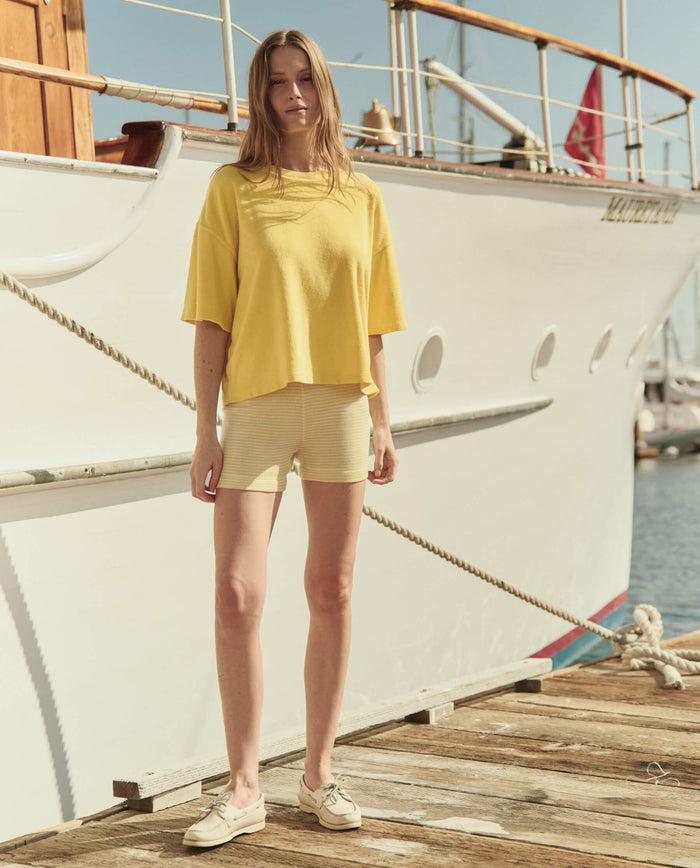 A woman wears THE GREAT. SU26 The Micro Terry Track Crew -- Margarita and light shorts as she stands on a wooden dock before a white boat on a sunny day.