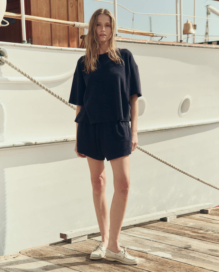 A woman in THE GREAT. SU26 The Micro Terry Track Crew — Navy, with matching shorts, stands on a wooden dock next to a white boat, looking at the camera.