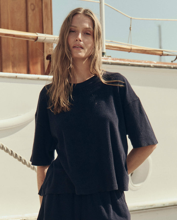 A woman with long hair stands outdoors in front of a boat, wearing THE GREAT. SU26 The Micro Terry Track Crew in navy.