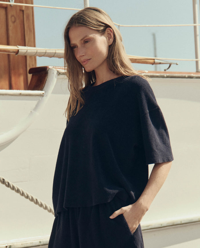 A woman with long hair stands outdoors near a boat, wearing THE GREAT. SU26’s The Micro Terry Track Crew in Navy, one hand in her pocket and looking slightly downward.