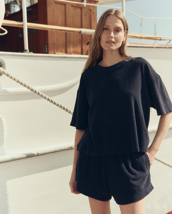 A woman stands outdoors on a boat, wearing THE GREAT. SU26 The Micro Terry Track Crew in navy and matching shorts, with one hand in her pocket.