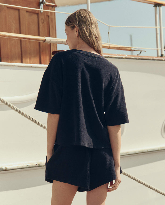 A person with long hair, wearing THE GREAT. SU26 The Micro Terry Track Crew in navy, stands on a boat deck facing away from the camera.