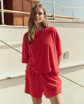A woman stands by a boat, wearing THE GREAT. SU26 The Micro Terry Track Crew in Pimento with matching red shorts.