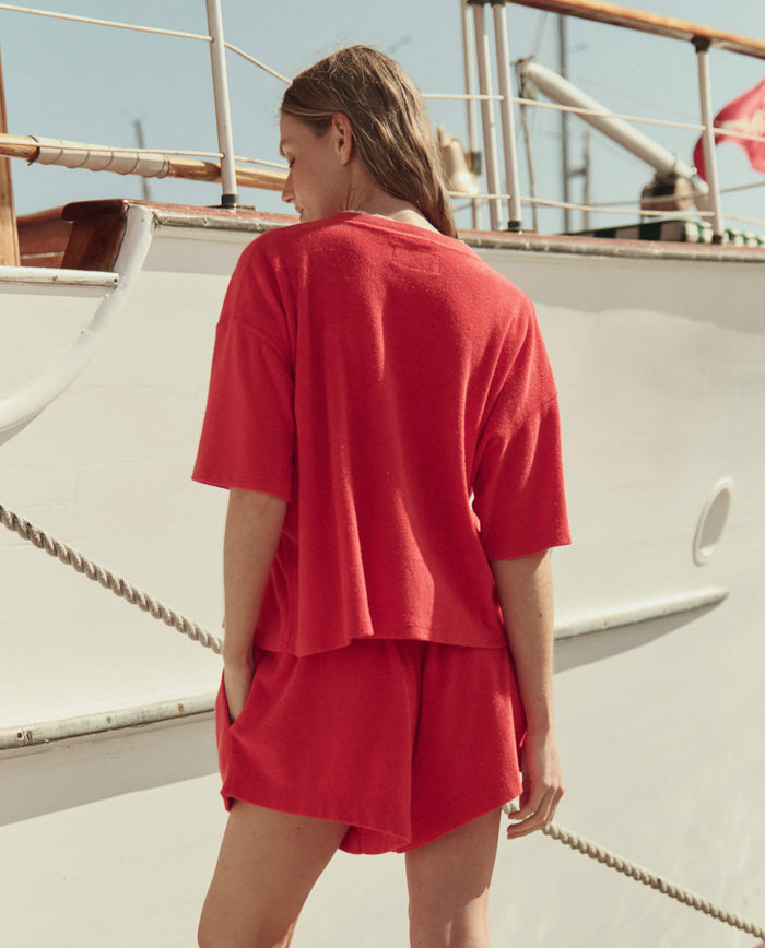 A woman in THE GREAT. SU26 Micro Terry Track Crew set in Pimento stands with her back to the camera by a white boat, channeling a relaxed track crew vibe.