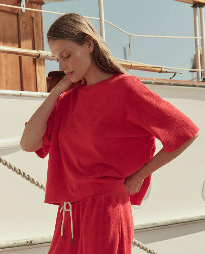 Wearing THE GREAT. SU26's Micro Terry Track Crew in Pimento, a woman stands by a boat, dressed in the loose-fitting red top and matching pants, with one hand on her head as she gazes downward.