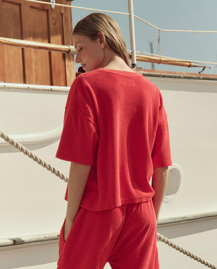 A person stands outdoors near a boat, hands in pockets, wearing THE GREAT. SU26's The Micro Terry Track Crew in Pimento, with matching pants, facing away from the camera.
